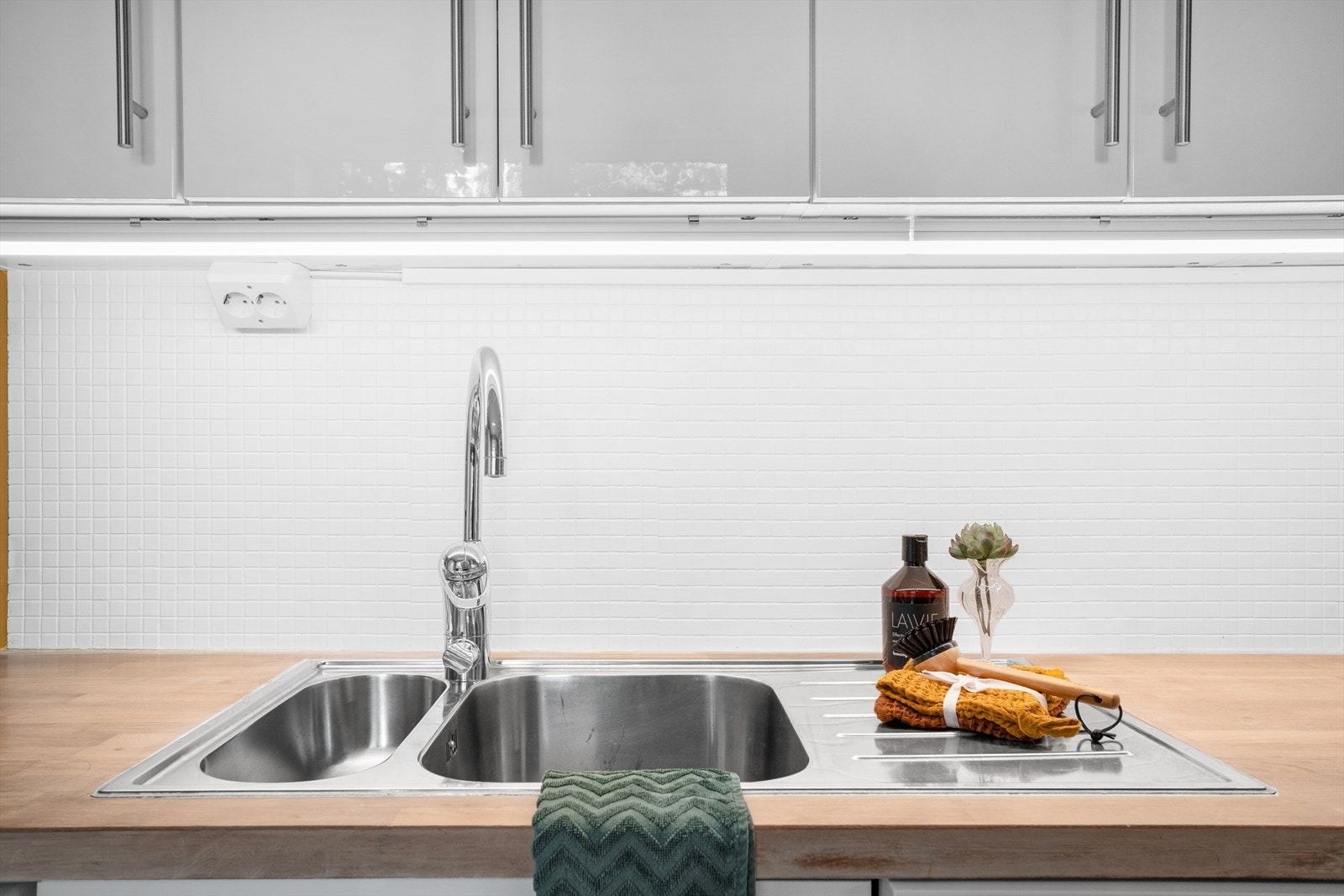 Mosaikkfliser på vegg mellom benk og overskap. Heltre benkeplate med nedfelt kum i stål. Galleribilde