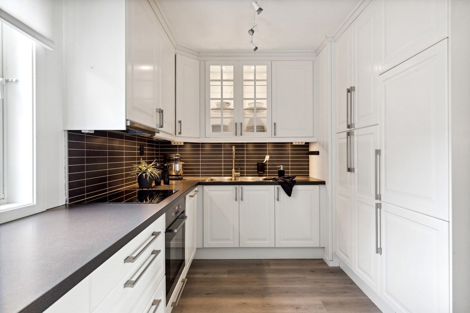 Den takhøye innredningen har hvite, profilerte fronter, vitrineskap og mørk laminatbenkeplate med dobbel oppvaskkum. Galleribilde