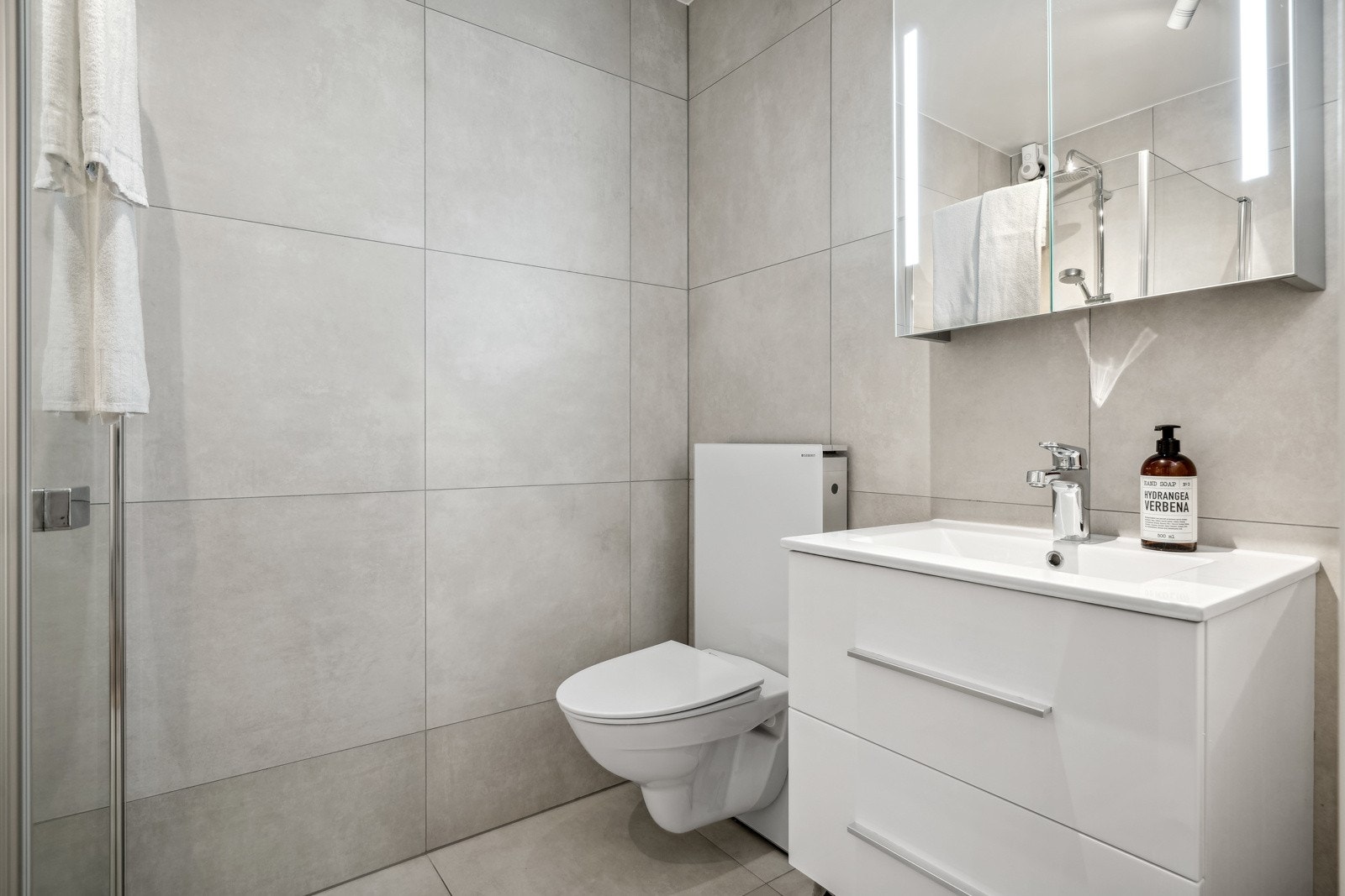 Badet er flislagt og framstår rålekkert med store fliser, vegghengt toalett og innredning med servant, skuffer og speilskap med belysning. Galleribilde