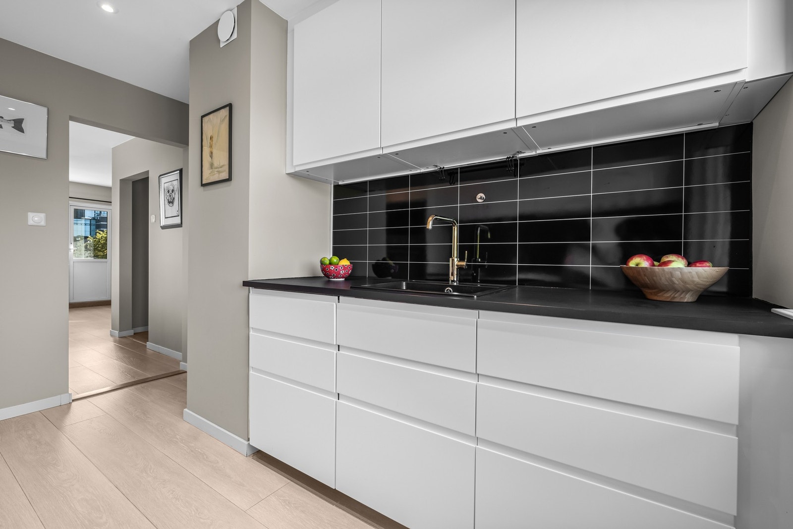 Glatte hvite fronter og vitrineskap, sort benkeplate nedfelt kum i svart kompositt, sorte flislignende veggplater på vegg samt lys under overskap. Galleribilde
