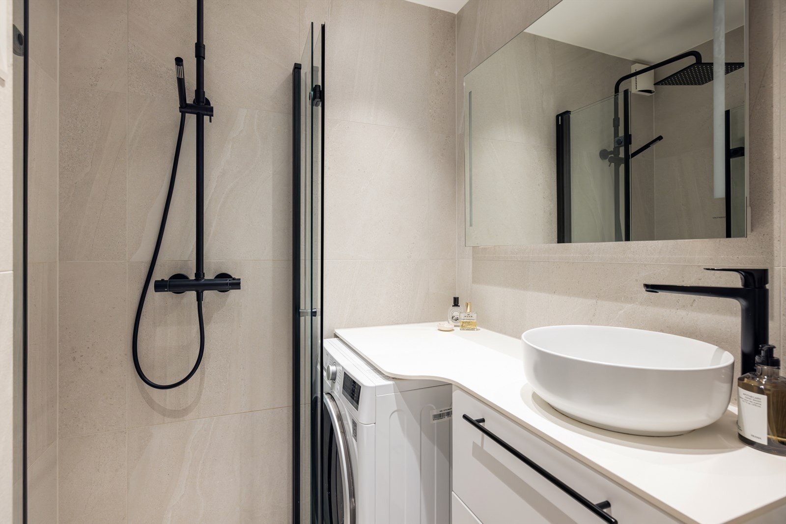 Innredningen inkluderer en rund, stilren servant montert på perfekt tilpasset benkeplate med skuffeinnredning under, speil med belysning, vegghengt toalett og dusjhjørne med dusjvegger i herdet glass. Opplegg for vaskemaskin. Galleribilde