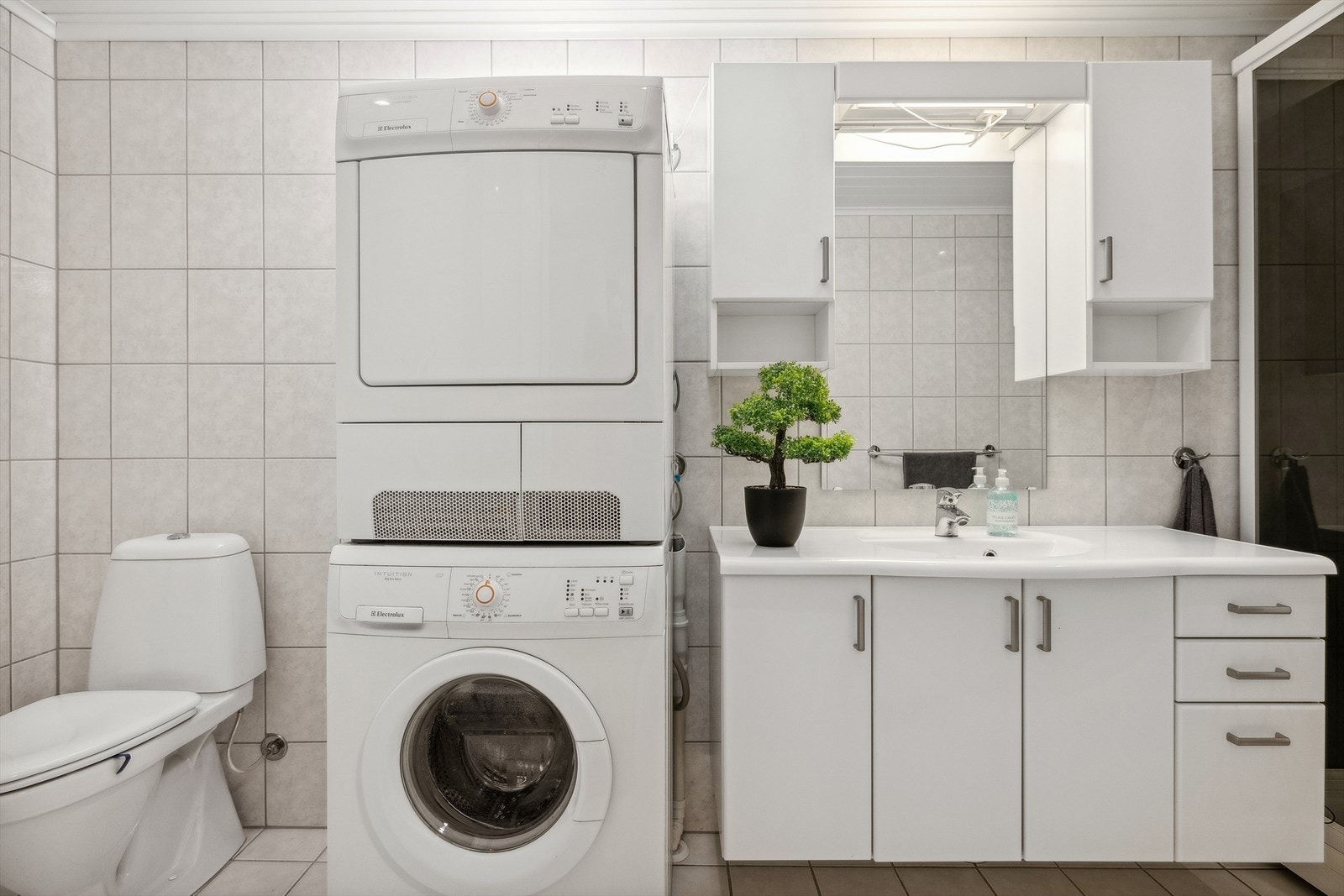Badet har flislagt gulv og vegger med MDF-panel i himling. Badet er innredet med toalett, opplegg til vaskemaskin, tørketrommel, servant med over og under-skap, speil og dusjkabinett Galleribilde