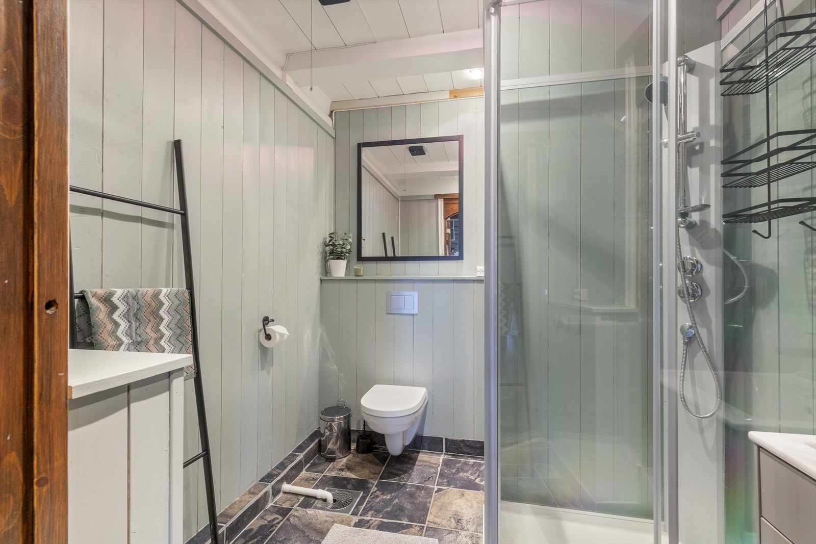 Bad 2 med veggmontert toalett, dusjkabinett med dører i glass og avtrekksvifte i himling. Det er flis på gulv med termostatstyrt varme. Galleribilde