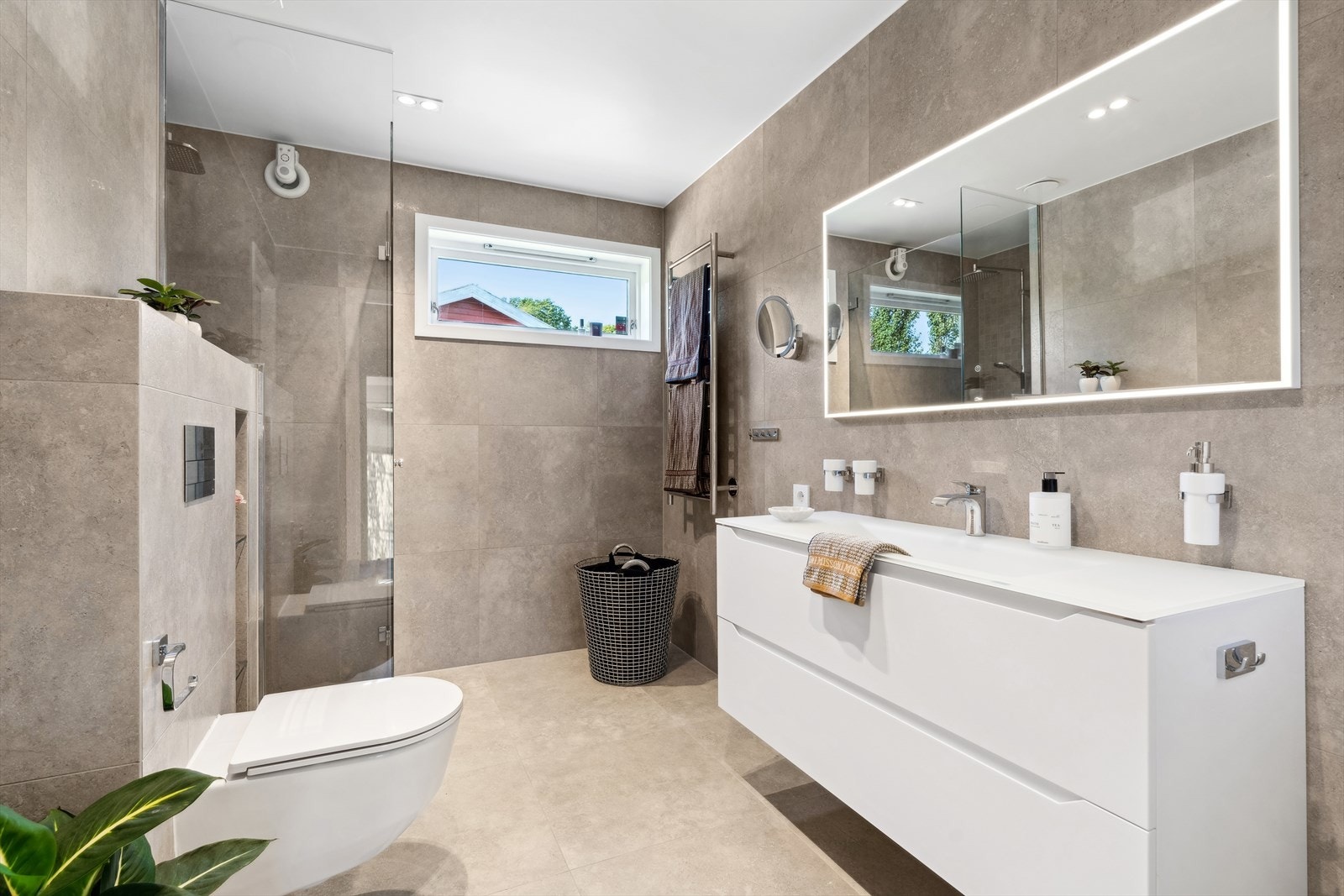 Rommet er innredet med nedfelt servant på hvit innredning med skuffer, speil, vegghengt toalett og dusjhjørne med glassvegger. Galleribilde