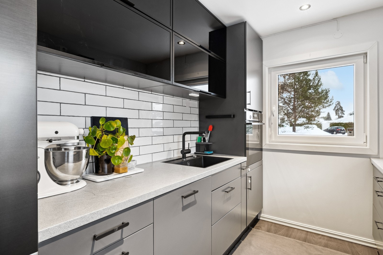 Pent kjøkken fra 2019 med glatte fronter, benkeplater i laminat, fliser over benkeplatene, oppvaskkum med ett-greps blandebatteri og ventilator over kokesonen. Galleribilde