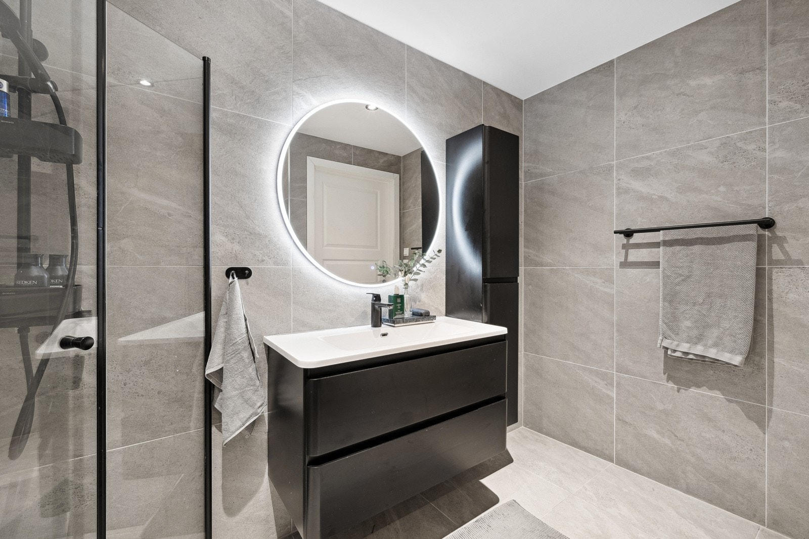 Mørk baderomsinnredning med servant, speil med belysning, høyskap, veggmontert toalett og dusjhjørne med regnfallsdusj og innfellbare glassdører for optimal romutnyttelse. Galleribilde