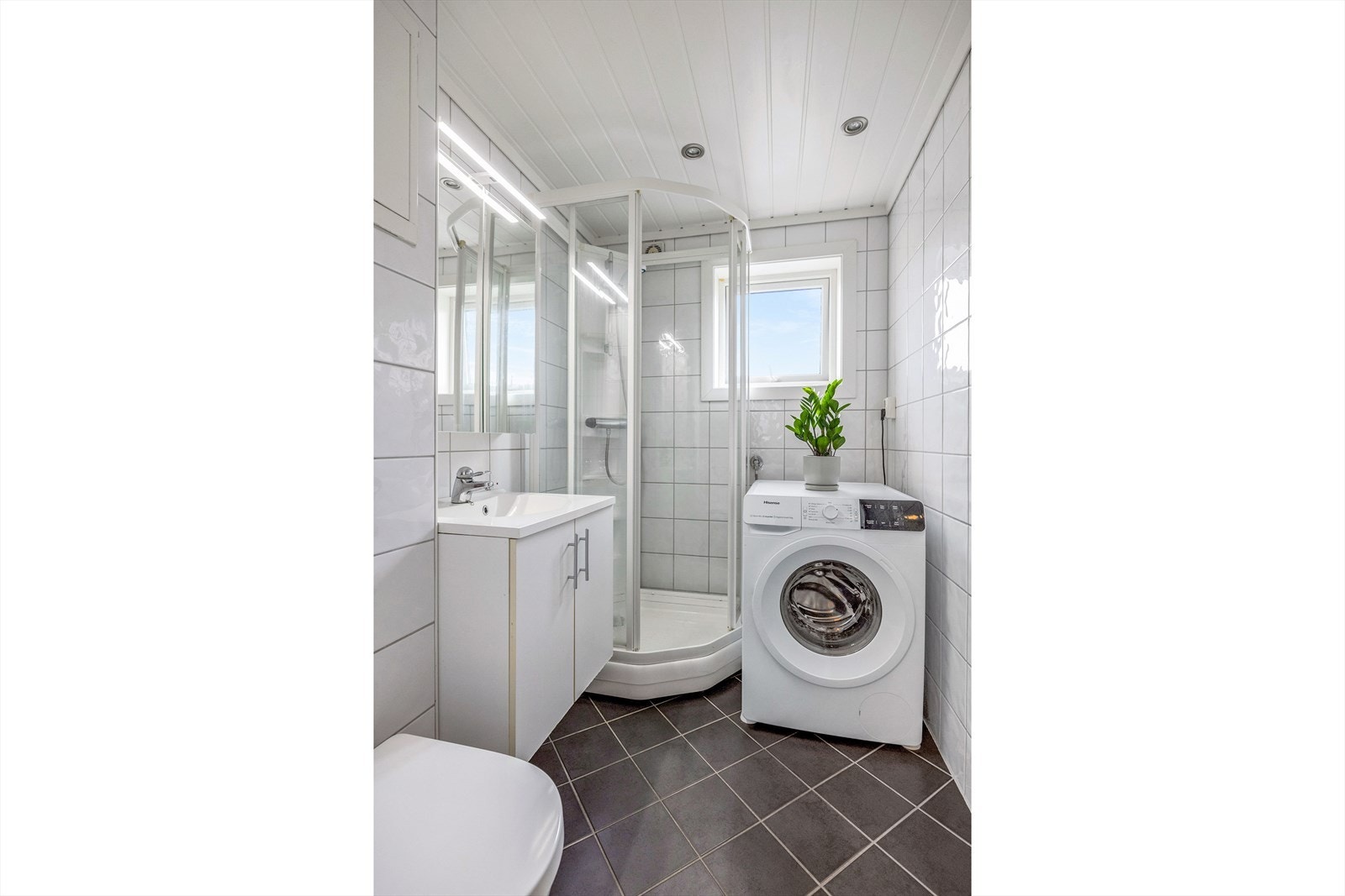 Pent flislagt baderom med varmekabler i gulv. Badet er utstyrt med servant over skapinnrending, overliggende speil med belysning, vegghengt toalett, dusjkabinett og opplegg til vaskemaskin Galleribilde