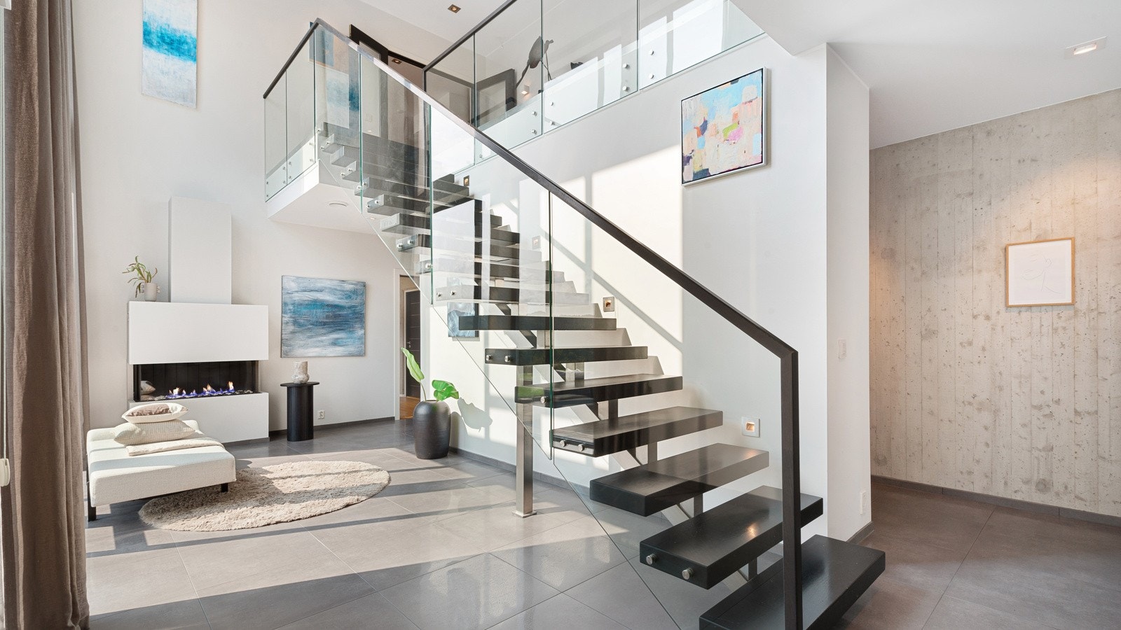 Nydelig og luftig hall med tiltalende gulvfliser, gasspeis og "flytende" trapp til hovedetasjen. Bak halvveggen er det godt med garderobeskap. Galleribilde