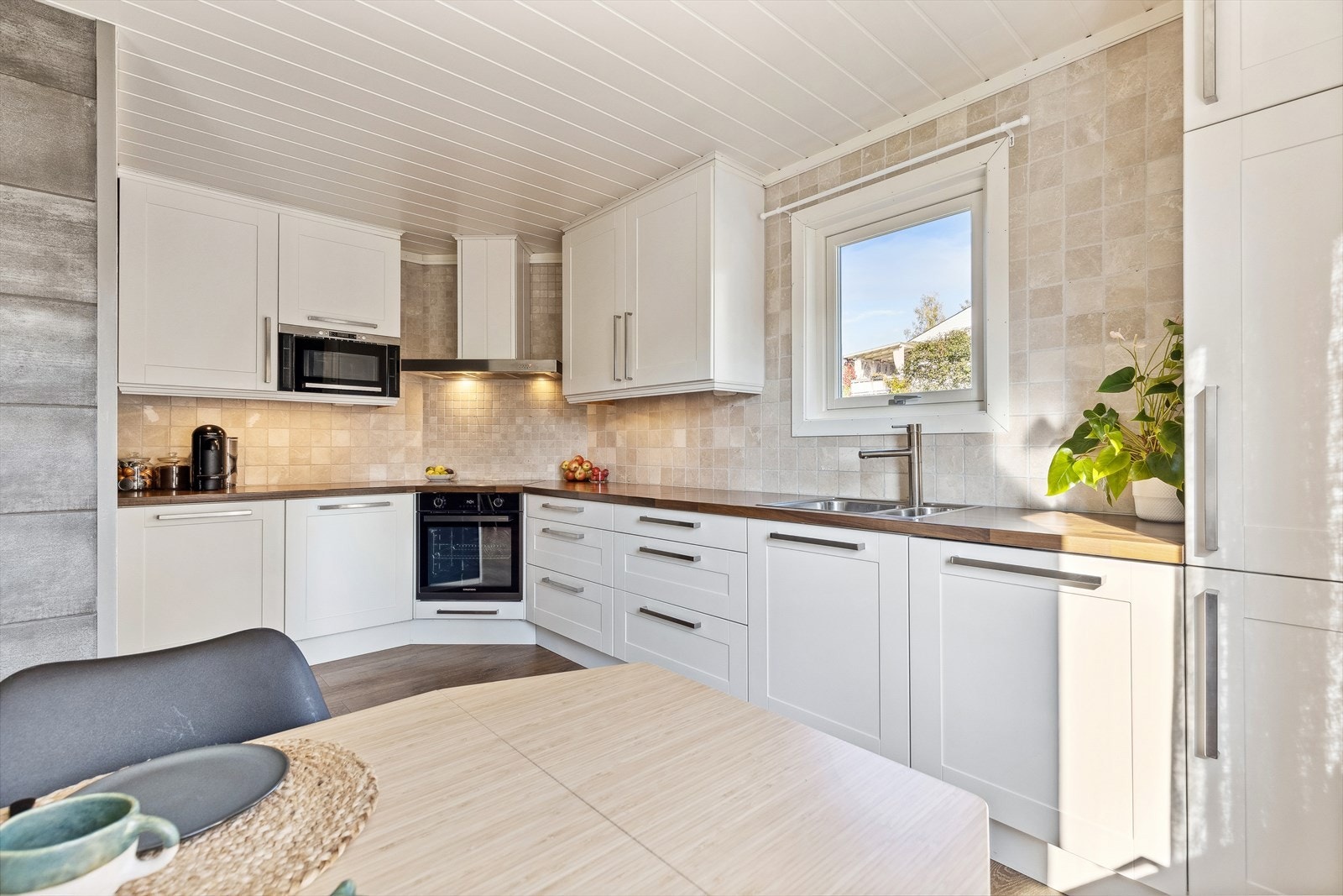 Innredningen har hvite, profilerte fronter og benkeplate med dobbel kjøkkenkum. Det er fliser over benkeplate og belysning under overskap. Galleribilde