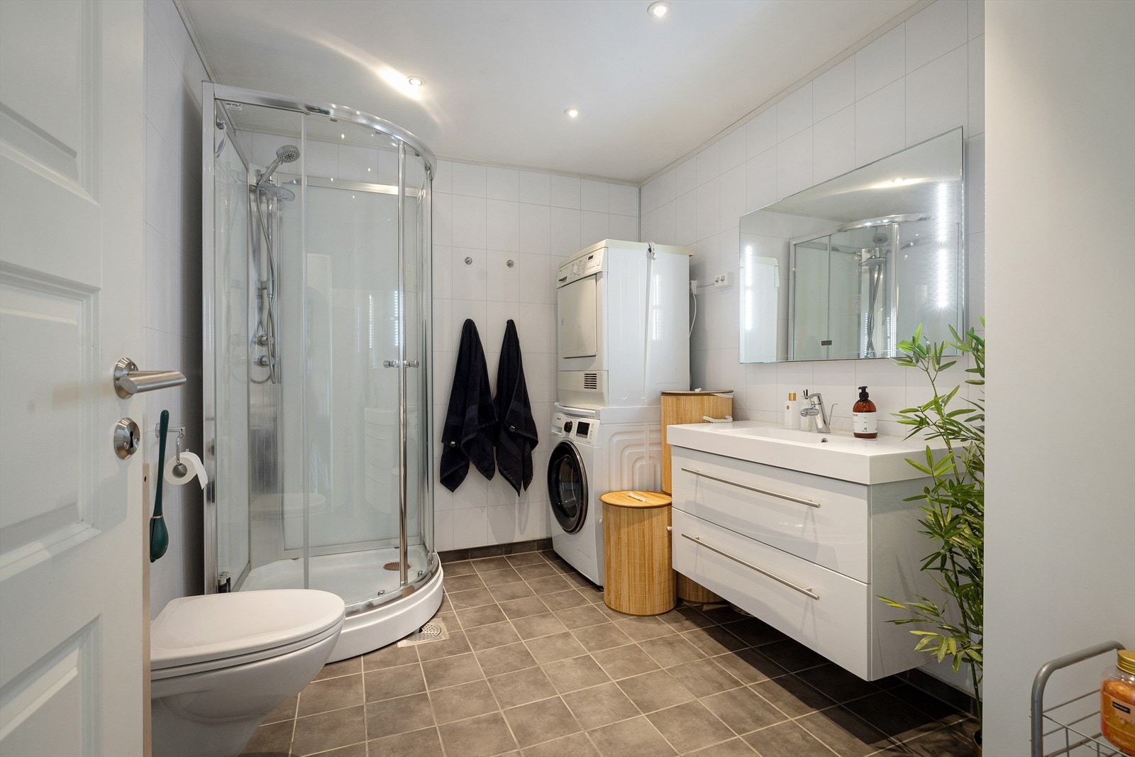 Her er det hvite våtromsplater i flisimitasjon på veggene, downlights i himlingen og sandfargede fliser på gulv med gulvvarme. Videre har badet et vegghengt toalett, et dusjkabinett og opplegg for vaskesøyle. Galleribilde