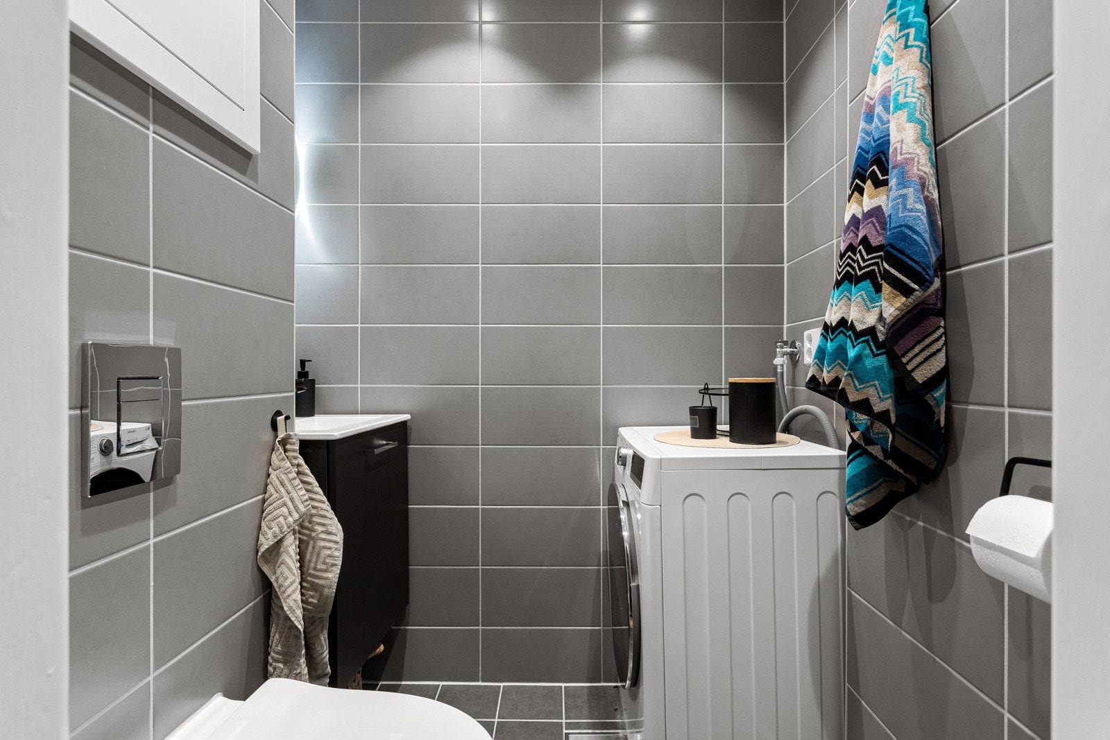 Toalettrommet er utstyrt med moderne servantskap med sort armatur, rundt speil med integrert belysning og vegghengt toalett. Galleribilde