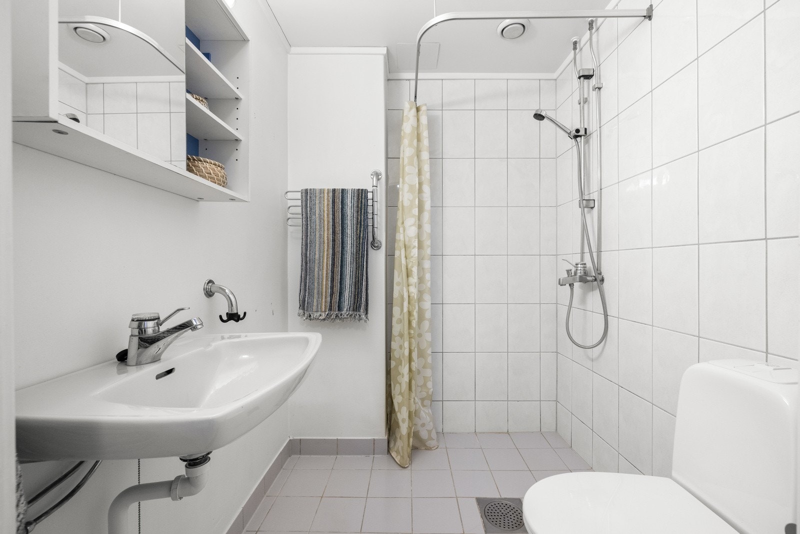 Badet er utstyrt med vegghengt servant med ett-greps armatur, speilskap med overbelysning og stikkontakt, dusjhjørne med forheng og vegghengt dusjarmatur tilkoblet hånddusj gulvstående toalett. Galleribilde