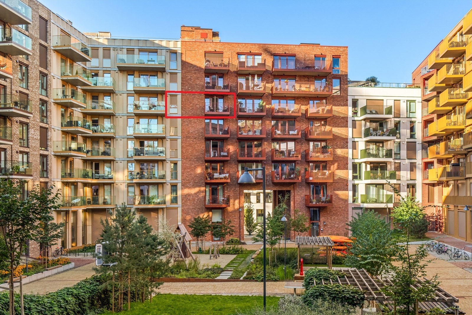 Pent opparbeidet bakgård med belegningssteinen, plen, trær, beplantning, lekeapparater, kajakkstativ og sykkelparkering. Galleribilde