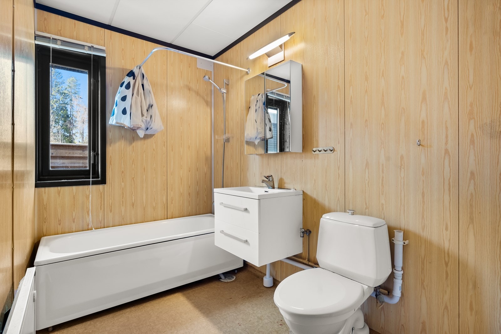 Baderom med våtromsbelegg på gulv og våtromsplater på veggene. Badekar med dusj, servant med speilskap og underliggende skuffeseksjoner og gulvstående toalett. Galleribilde