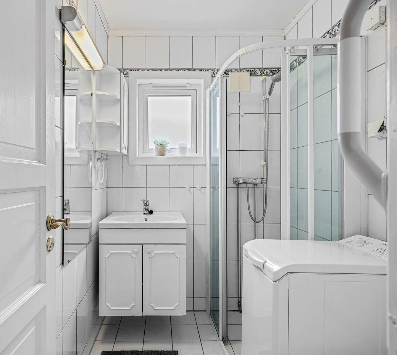 Flislagt bad med gulvvarme og opplegg for vaskemaskin. Innredningen består av et underskap/overskap, servant, speil og armatur med stikk og en dusjsone med skyvedører. Galleribilde