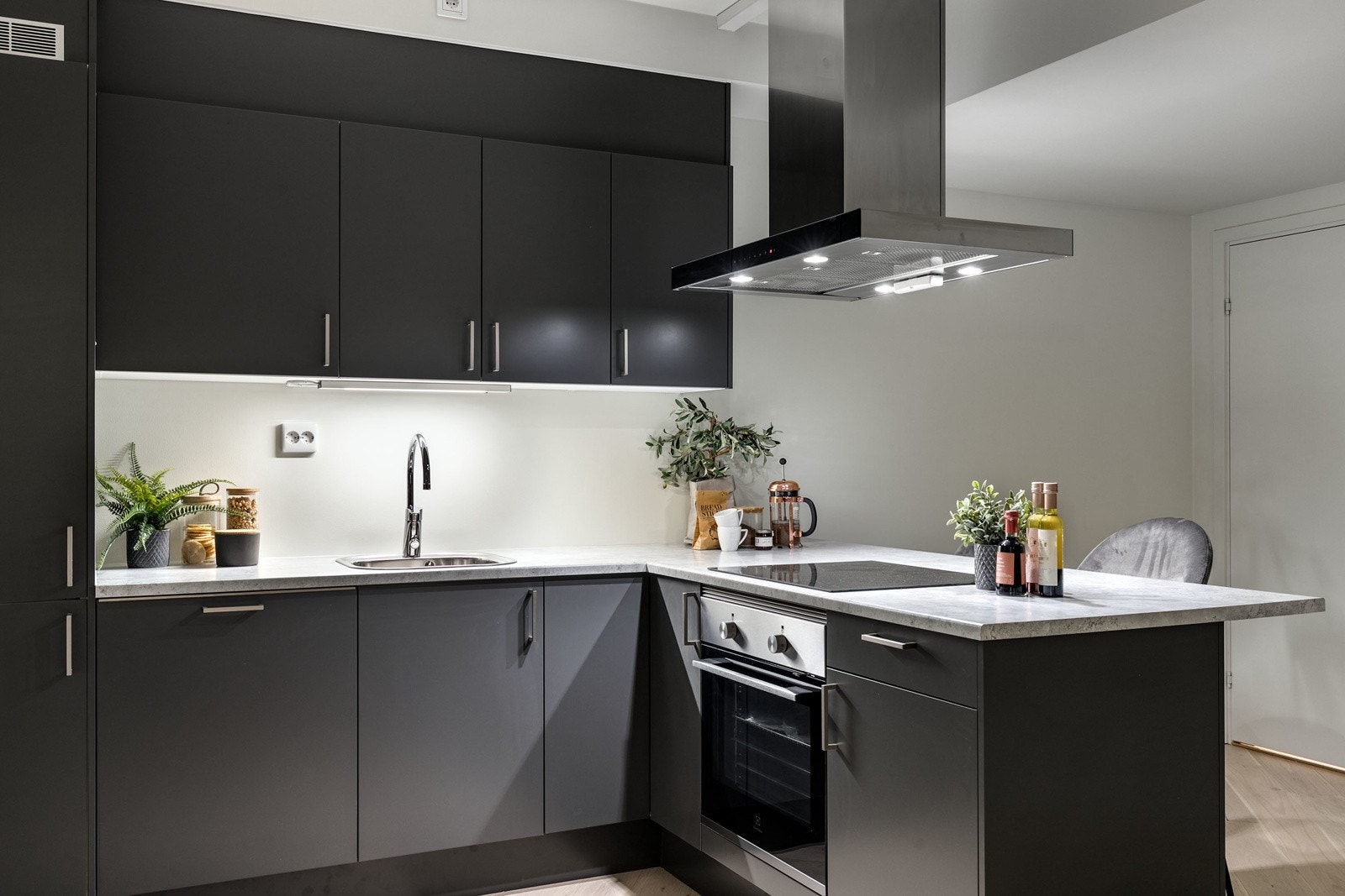 Stilrent og moderne kjøkken fra Sigdal i åpen løsning mot stuen. Innredning med mørke slette fronter med foring opp til tak. Laminert benkeplate i lys utførelse med nedfelt oppvaskkum i rustfritt stål. Det er montert belysning under overskap. Galleribilde