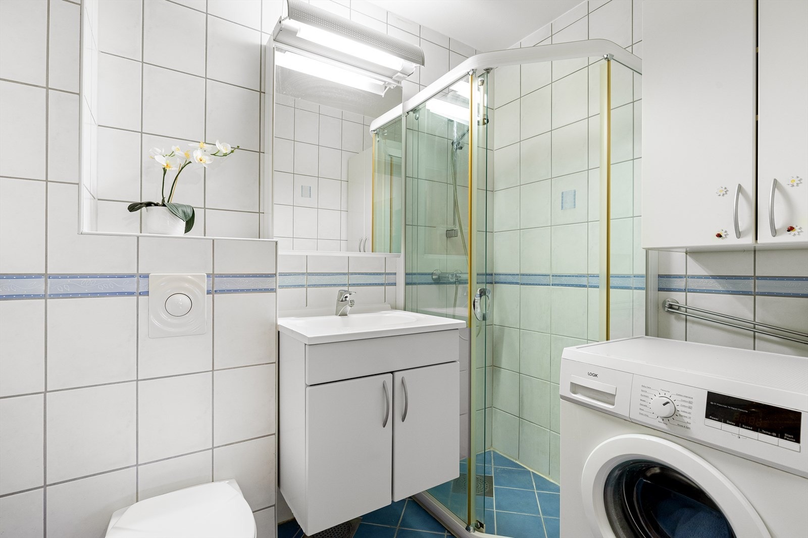 Badet inneholder veggmontert toalett, servant med baderomsinnredning, praktisk speilskap med belysning, dusjhjørnet på flis og opplegg til vaskemaskin. Galleribilde