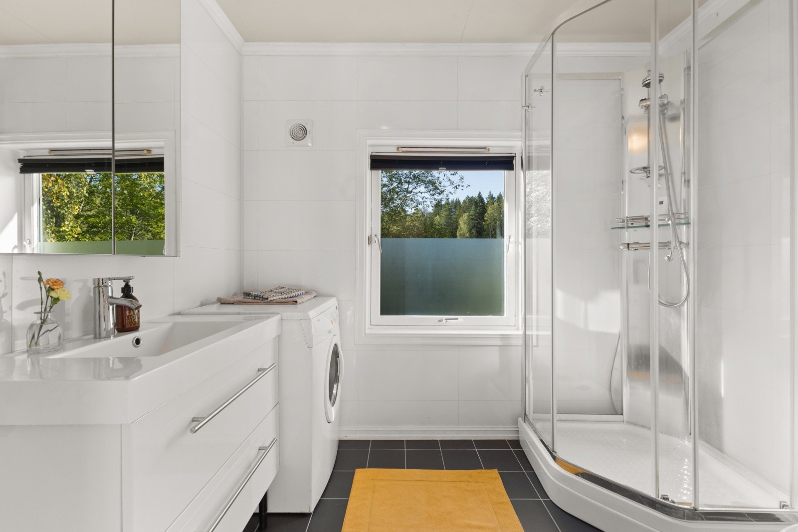 Baderom med flislagt gulv med varme og baderomsplater på vegg. Badet inneholder gulvstående servantskap, ovenpåliggende servant, speilskap og stikkontakt på vegg over servant. Galleribilde