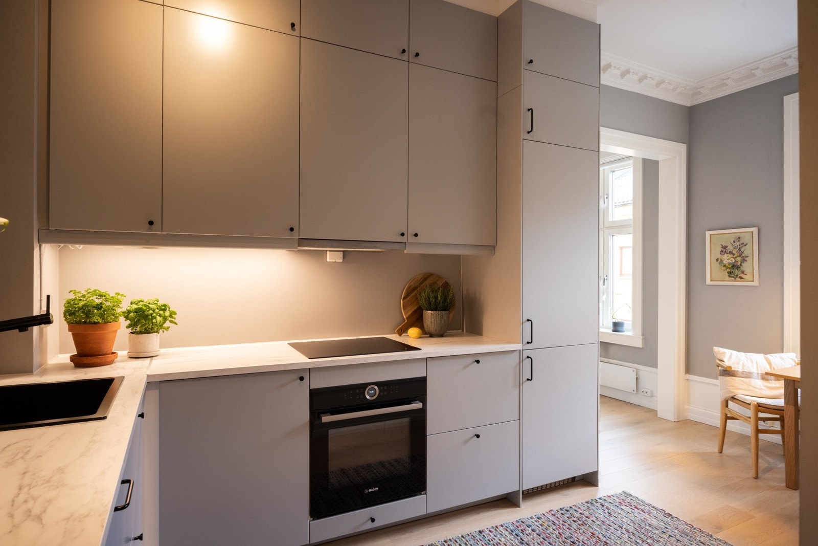 Pen kjøkkeninnredning med slette overflater i en delikat gråtone. Kjøkkenet har rikelig med oppbevaringsplass, hvor overskapene strekker seg helt opp til taket - perfekt for ekstra lagringsplass og et ryddig uttrykk. Galleribilde