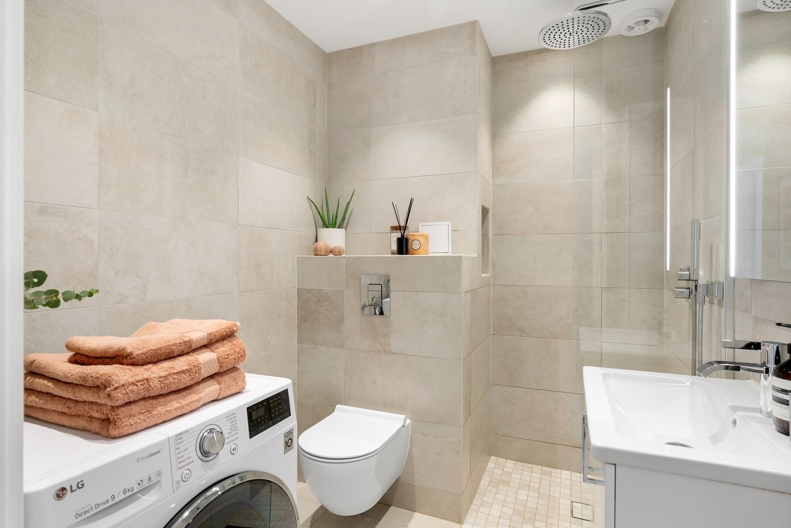 Badet er utstyrt med dusjhjørne med dusjskillevegg i herdet glass, helservant med underskap, speilskap med LED lys over servant, høyskap, vegghengt toalett og opplegg for vaskemaskin/tørketrommel. Galleribilde