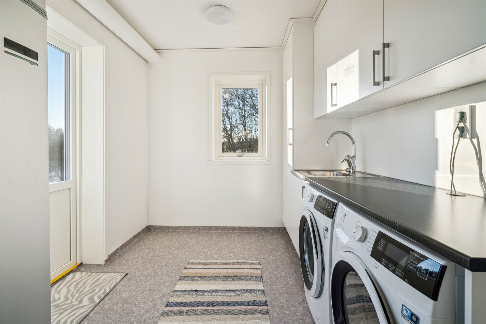 Vaskerommet har lysmalte vegger og grått belegg med oppbrett på gulvet. Rommet er innredet med vegghengt innredning med skap og benkeplate. Nedfelt vaskekum og ett-greps blandebatteri. Galleribilde