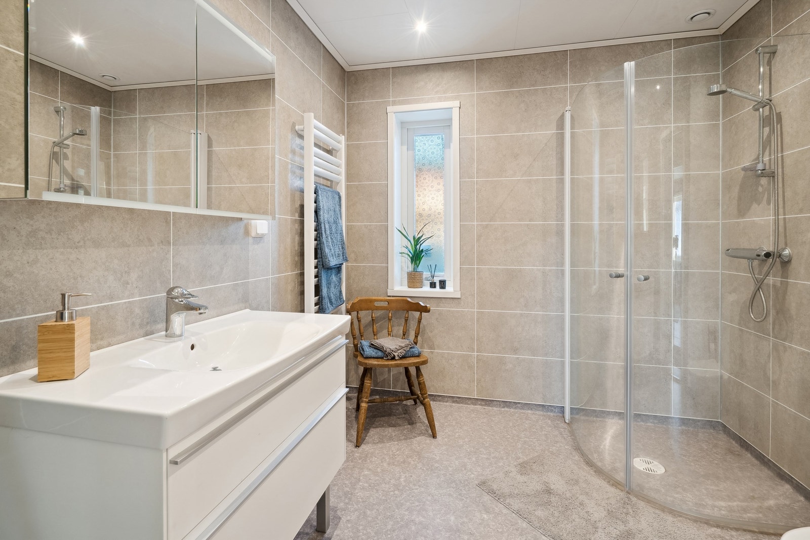 Baderommet er innredet med heldekkende servant i hvit skuffeinnredning, speilskap, dusjhjørne med armatur montert på vegg og dusjvegger og gulvmontert toalett. Balansert ventilasjon i rommet. Galleribilde