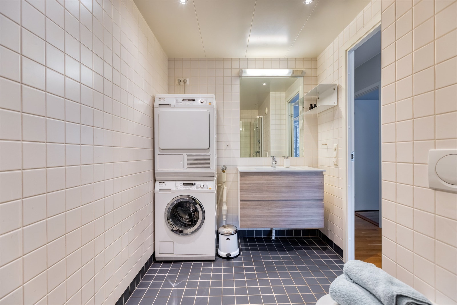 Flislagt pent bad med servantskap med skuffer, vegghengt toalett, dusjhjørne og opplegg for vaskemaskin. Galleribilde