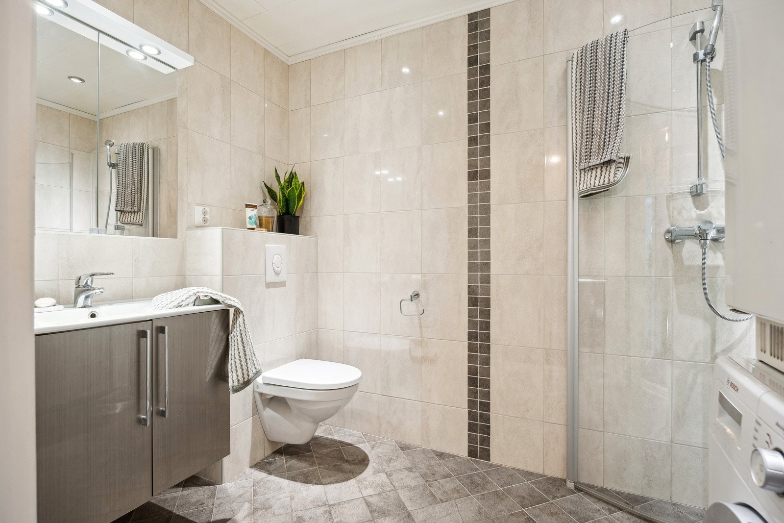 Flislagt baderom utstyrt med vegghengt servantinnredning, speilskap, dusjhørne og vegghengt toalett. Galleribilde