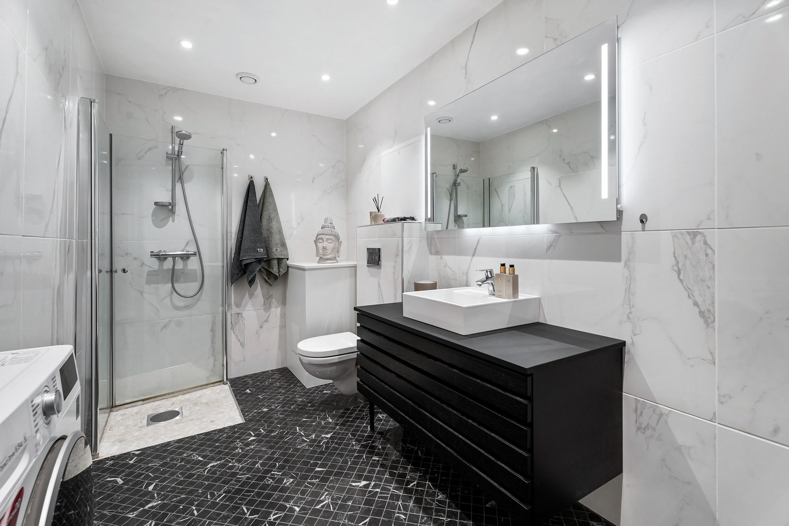 Badet er utstyrt med vegghengt servantinnredning med skuffer, ovenpåliggende servant med ett-greps armatur, speil med sidelys og vegghengt toalett. Galleribilde