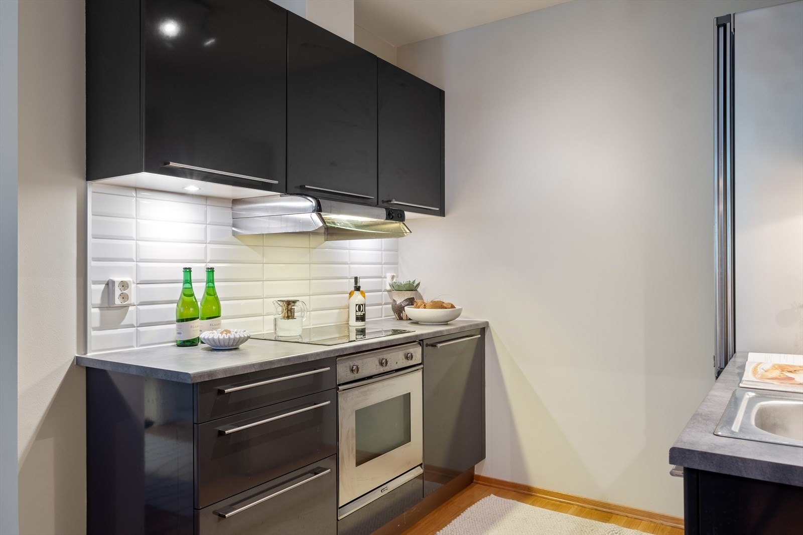 Kjøkkeninnredning med glatte fronter, laminat benkeplate med nedfelt vaskekum. Fliser og belysning over benkeplaten. Galleribilde