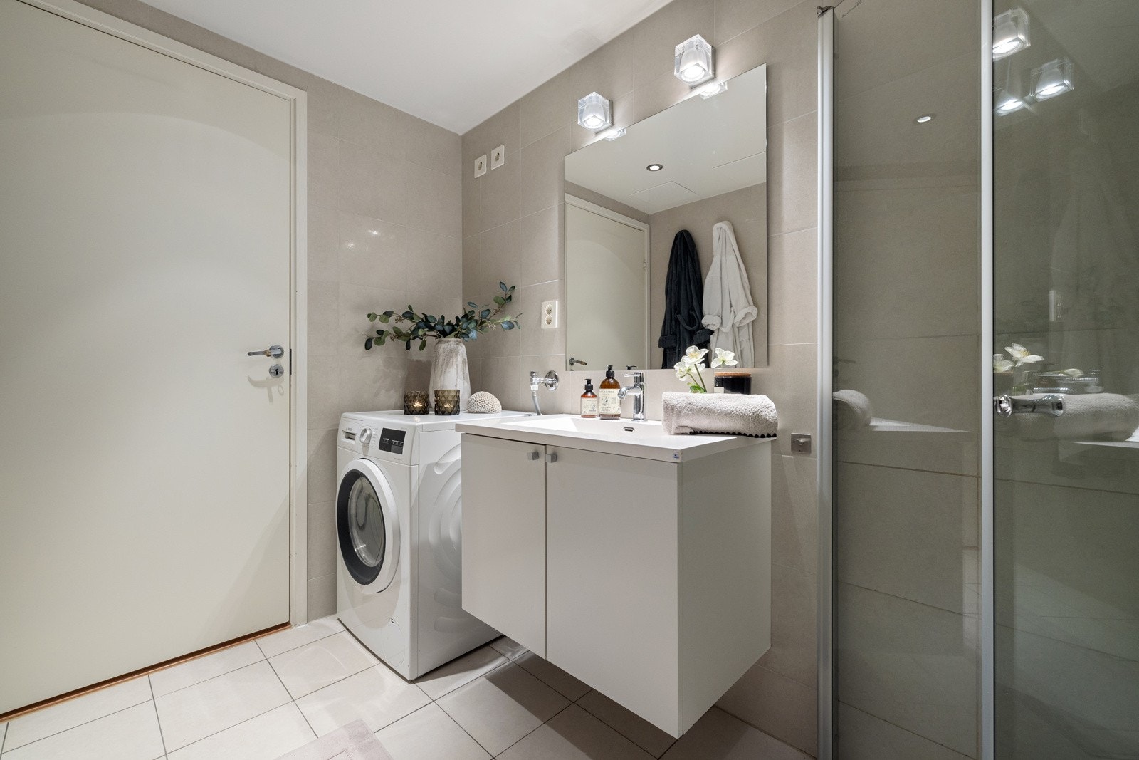 Badet er innredet med en klassisk baderomsløsning som inkluderer lyse fronter, en heldekkende servant, speil med integrert belysning, vegghengt toalett og en hjørnedusj med glassdører. Galleribilde