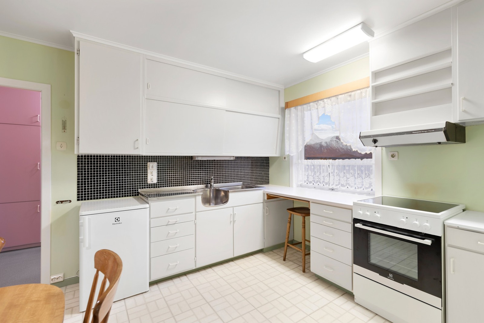 Kjøkkenet har montert en eldre, L-formet innredning med benkeskap og takhøye overskap. Opplegg for frittstående kjøleskap og komfyr Galleribilde