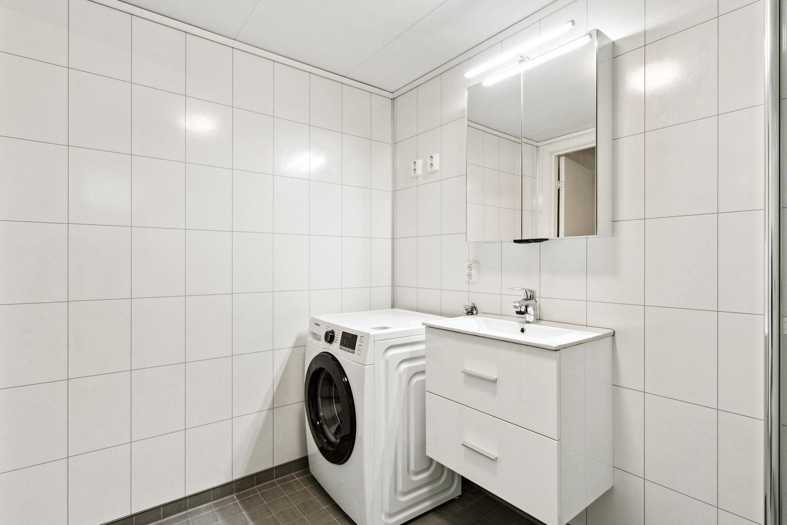 Delikat bad med grå gulvfliser, hvite våtromsplater, opplegg for vaskemaskin og varmekabler. Galleribilde