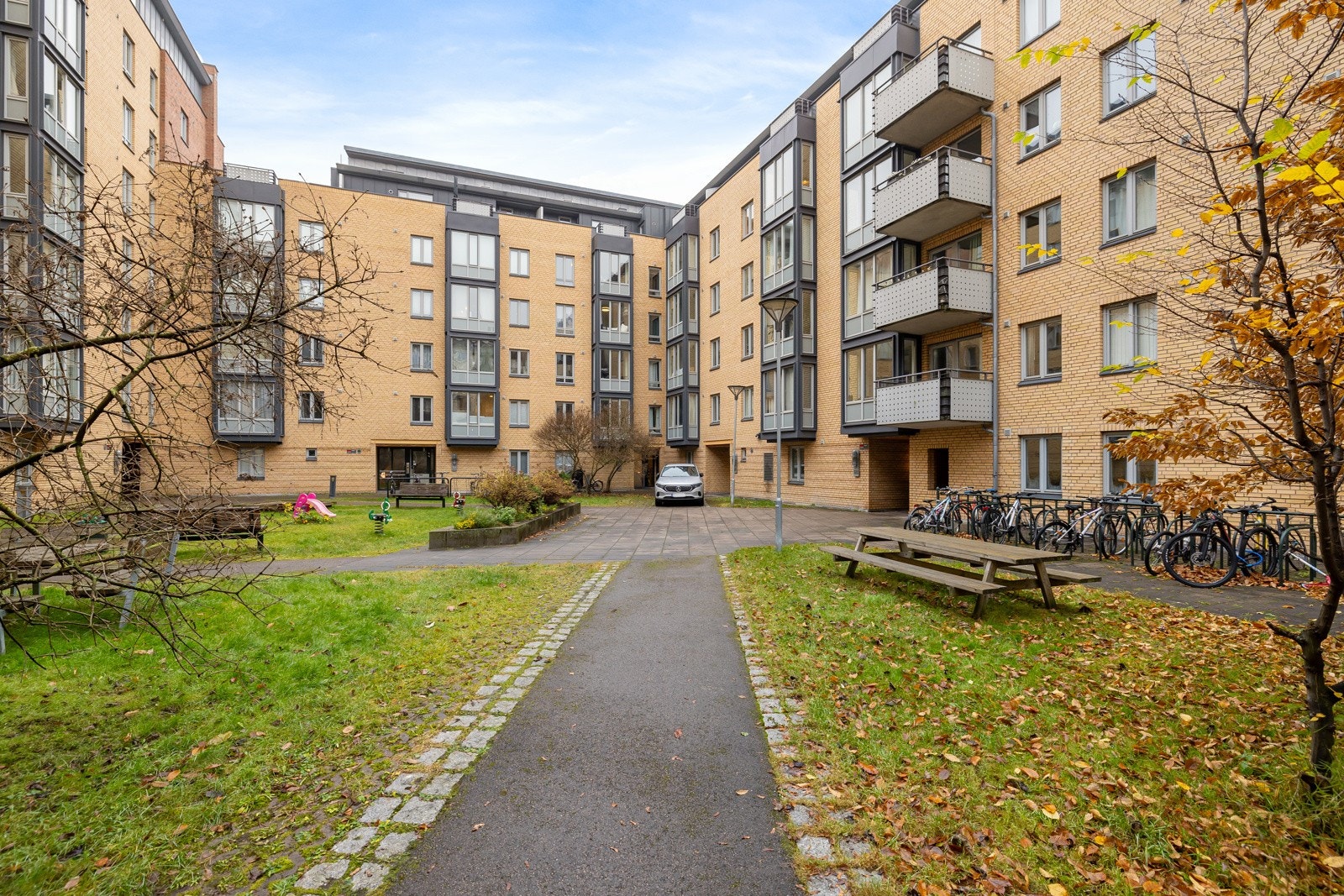 Pent opparbeidet bakgård med sittegrupper, prydbukser, beplantning, internveier, sykkelstativ og lekestativ. Galleribilde