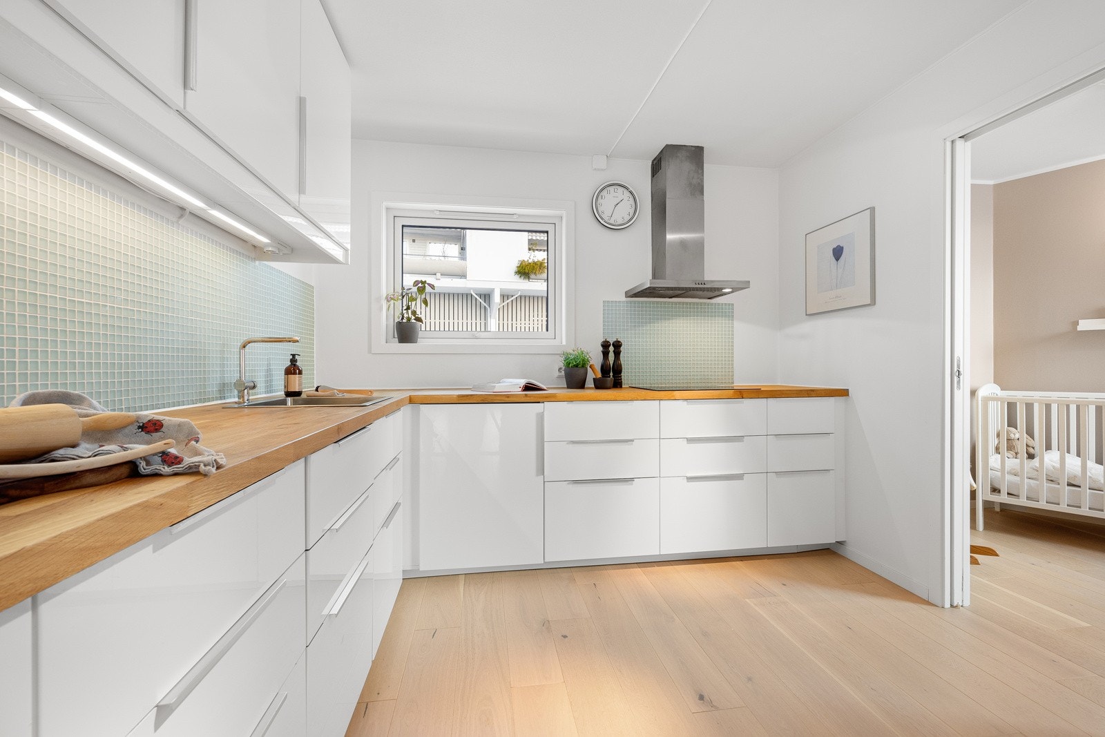 Innredningen har takhøye skap med hvite høyglansfronter, benkeplate i heltre og glassmosaikk som backsplash. Galleribilde