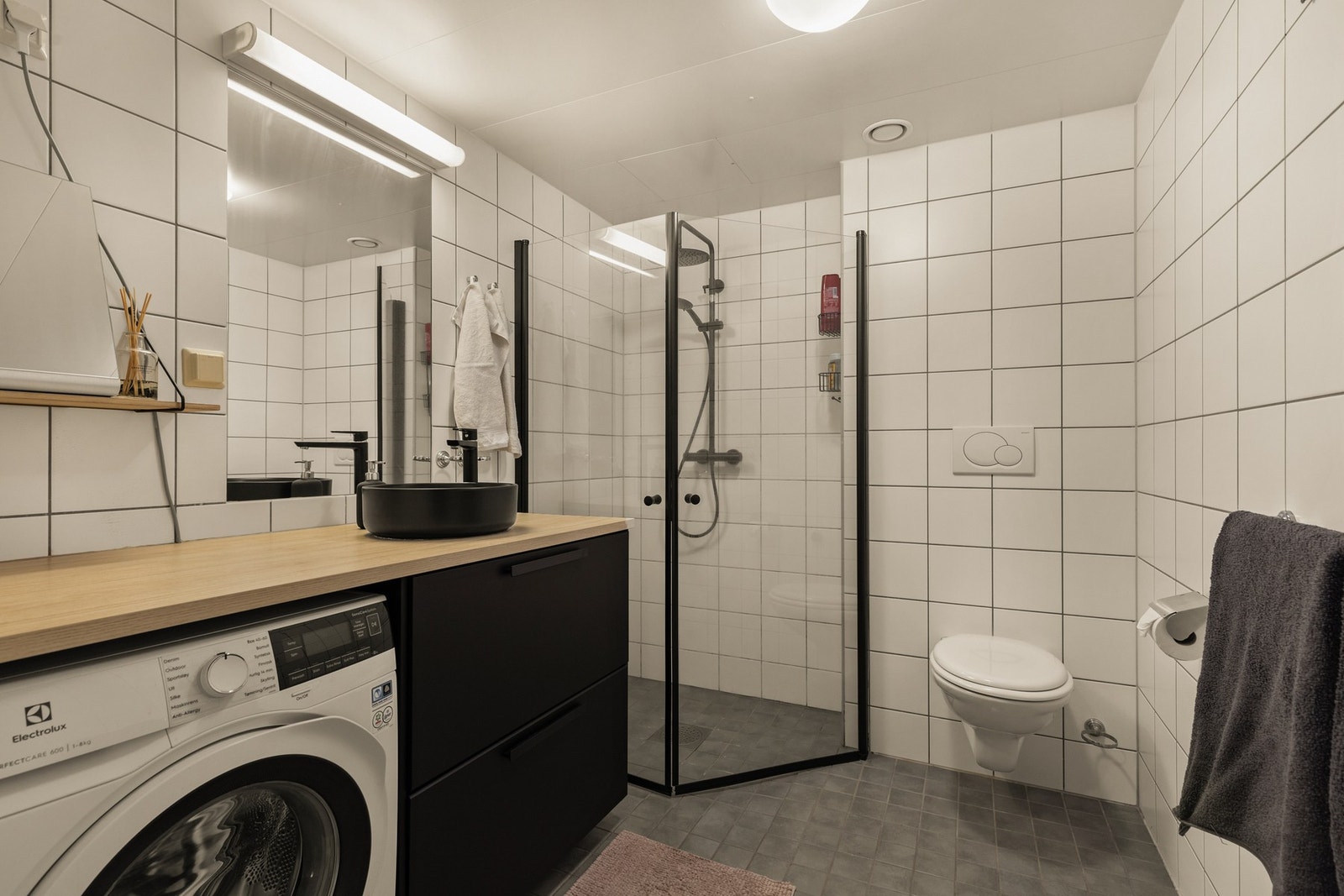 Badet har tidløse hvite fliser på vegg og grå fliser på gulv. Her får du pen baderomsinnredning med sorte detaljer og en lekker benk Galleribilde