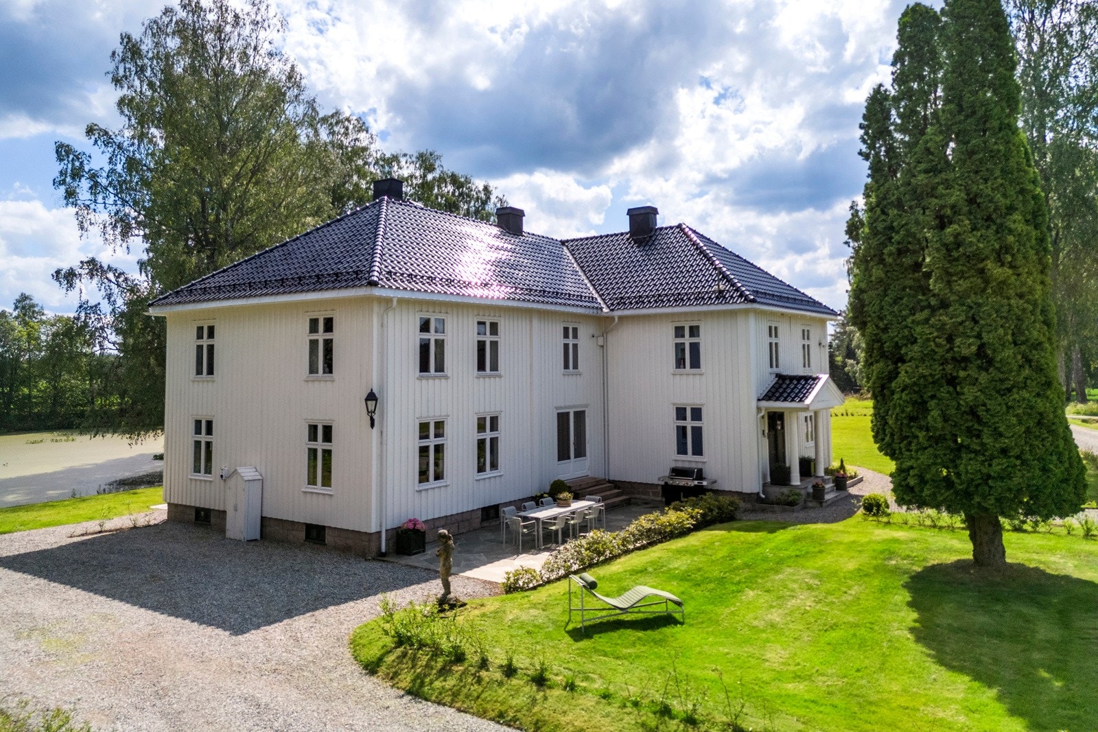 Den storslåtte hovedbygningen troner majestetisk på gården, omgitt av grønne marker og vakre gamle trær. Galleribilde