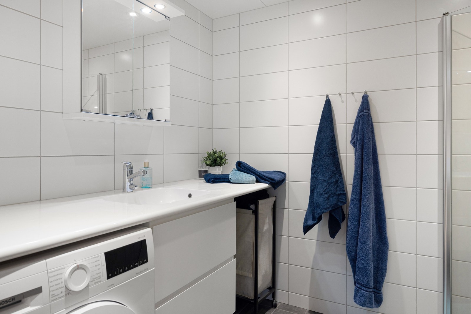 Den hvite innredningen har glatte skuffefronter og et speilskap med praktisk belysning over. Heldekkende benkeplate med servant gir et stilrent uttrykk, og det er plass til vaskemaskin under benkeplaten. Galleribilde