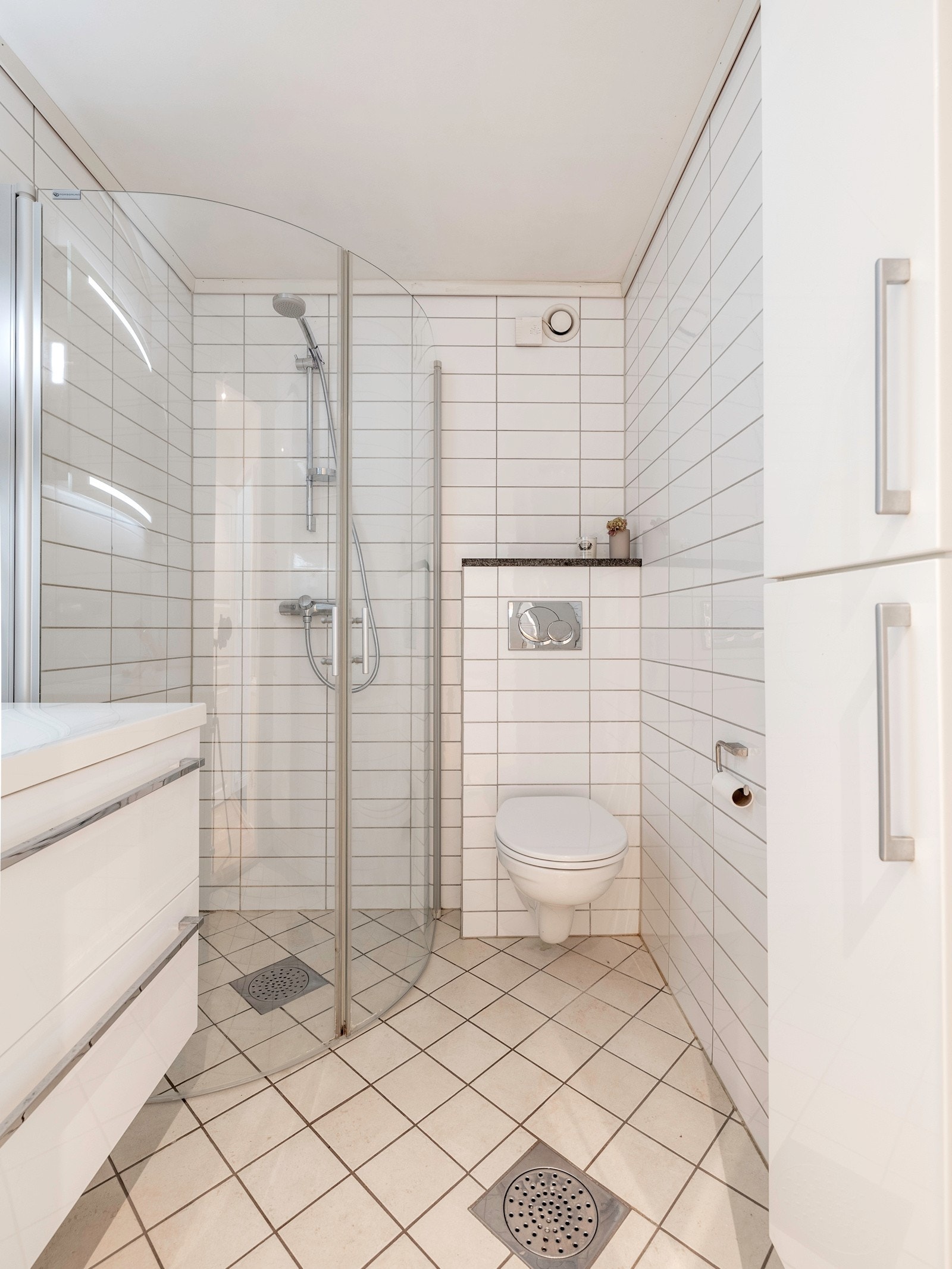Innredningen er i moderne stil med hvite, glatte skuffefronter, heldekkende servant, matchende høyskap og speil med integrert belysning. Badet har ellers et veggmontert toalett og dusjhjørne med buede, innfellbare glassdører. Galleribilde