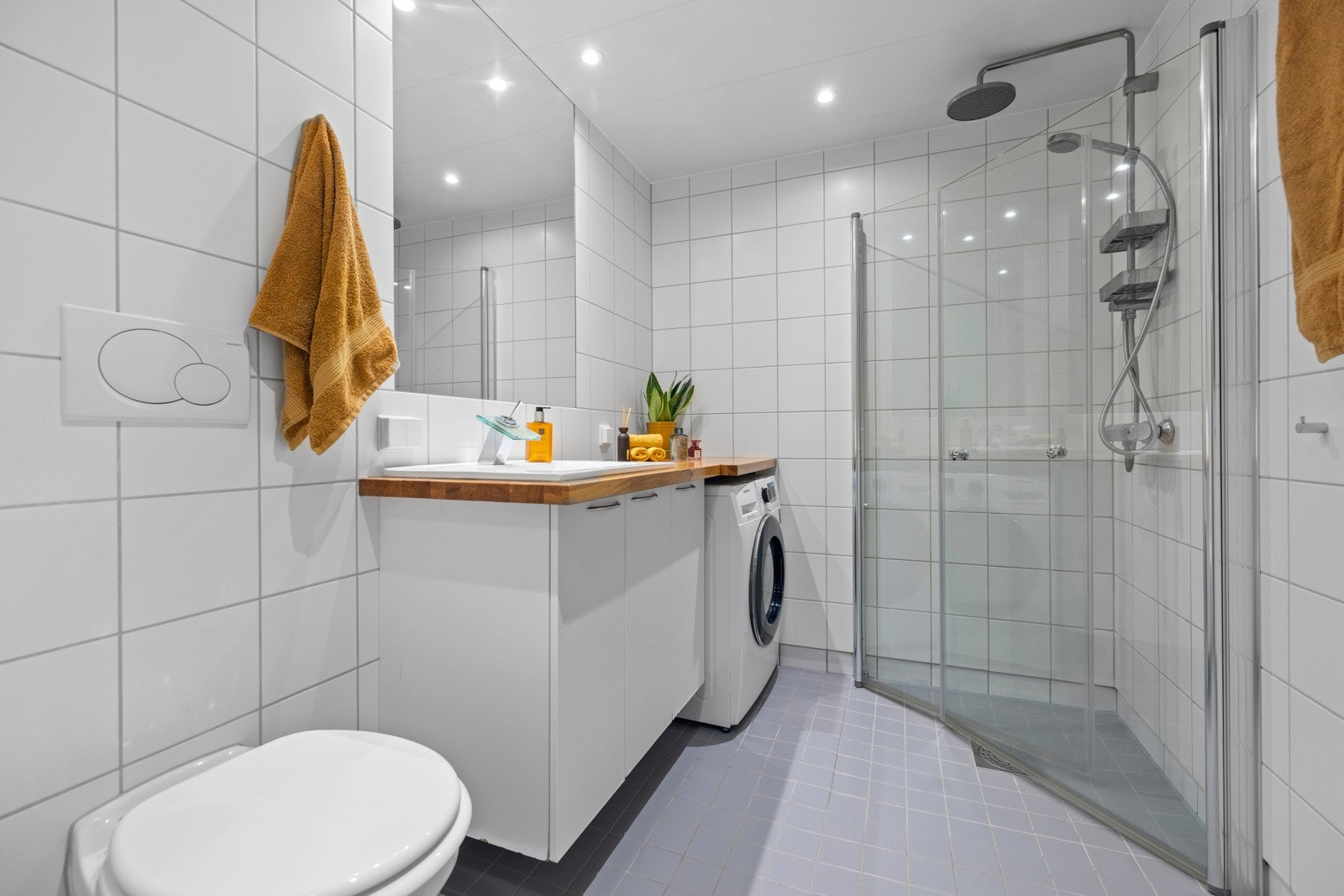 Flislagt baderom fra byggeår utført i lyse fliser med downlights i himling. Nedsenket dusjhjørne med innfellbare glassdører og regnfallsdusj og vegghengt toalett Galleribilde