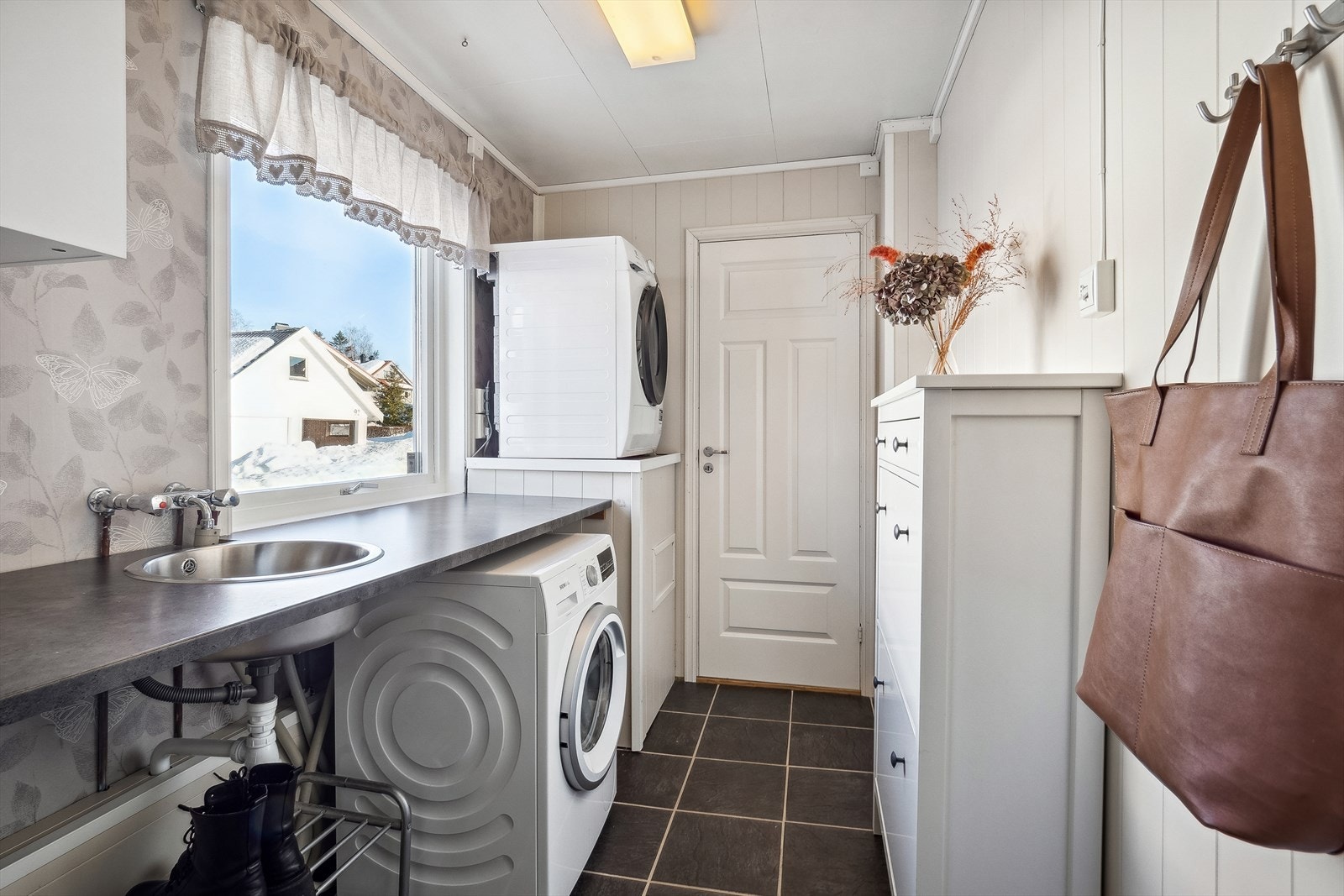 Vaskerom med panel/ tømmer på veggene og fliser på gulv med gulvvarme. Rommet har innredning med benkeplate med nedfelt servant, overskap og opplegg for vaskemaskin. Galleribilde