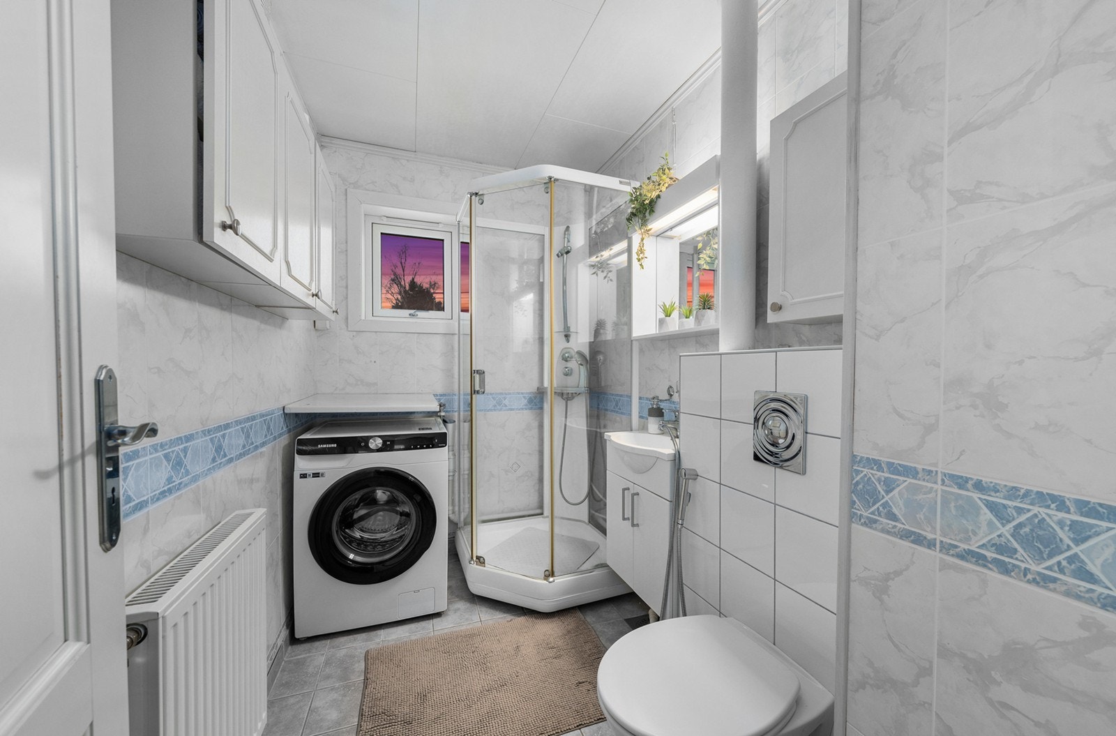 Bad med lys innredning med med nedfelt servant og speilinnredning med belysning. Badet er videre innredet med veggmontert toalett, dusjkabinett og opplegg for oppvaskmaskin. Galleribilde