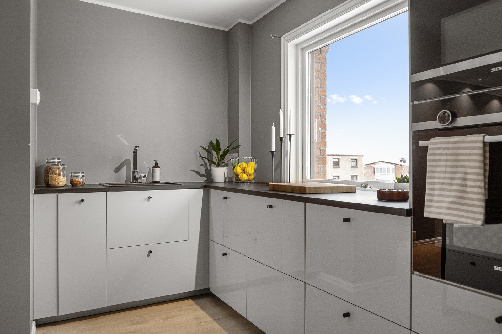 Mørk laminat benkeplate med nedfelt sort komposittvask og ett-greps armatur i krom. Kjøkkenet er utstyrt med integrerte hvitevarer fra Siemens, inkludert oppvaskmaskin, kjøl/frys, stekeovn, mikrobølgeovn i høyskap og platetopp Galleribilde