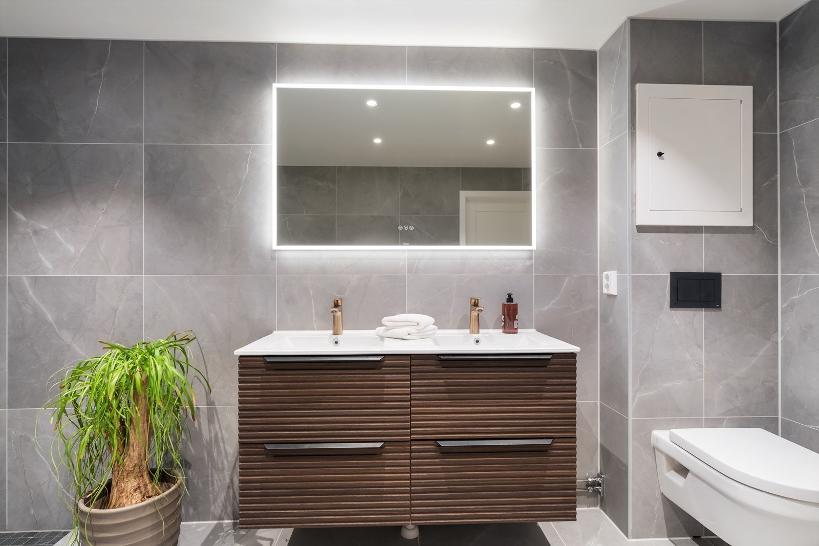 Badet er flislagt med grå fliser med marmormønster. Det er montert dobbelservant med romslig innredning og speil med innebygget belysning. Galleribilde