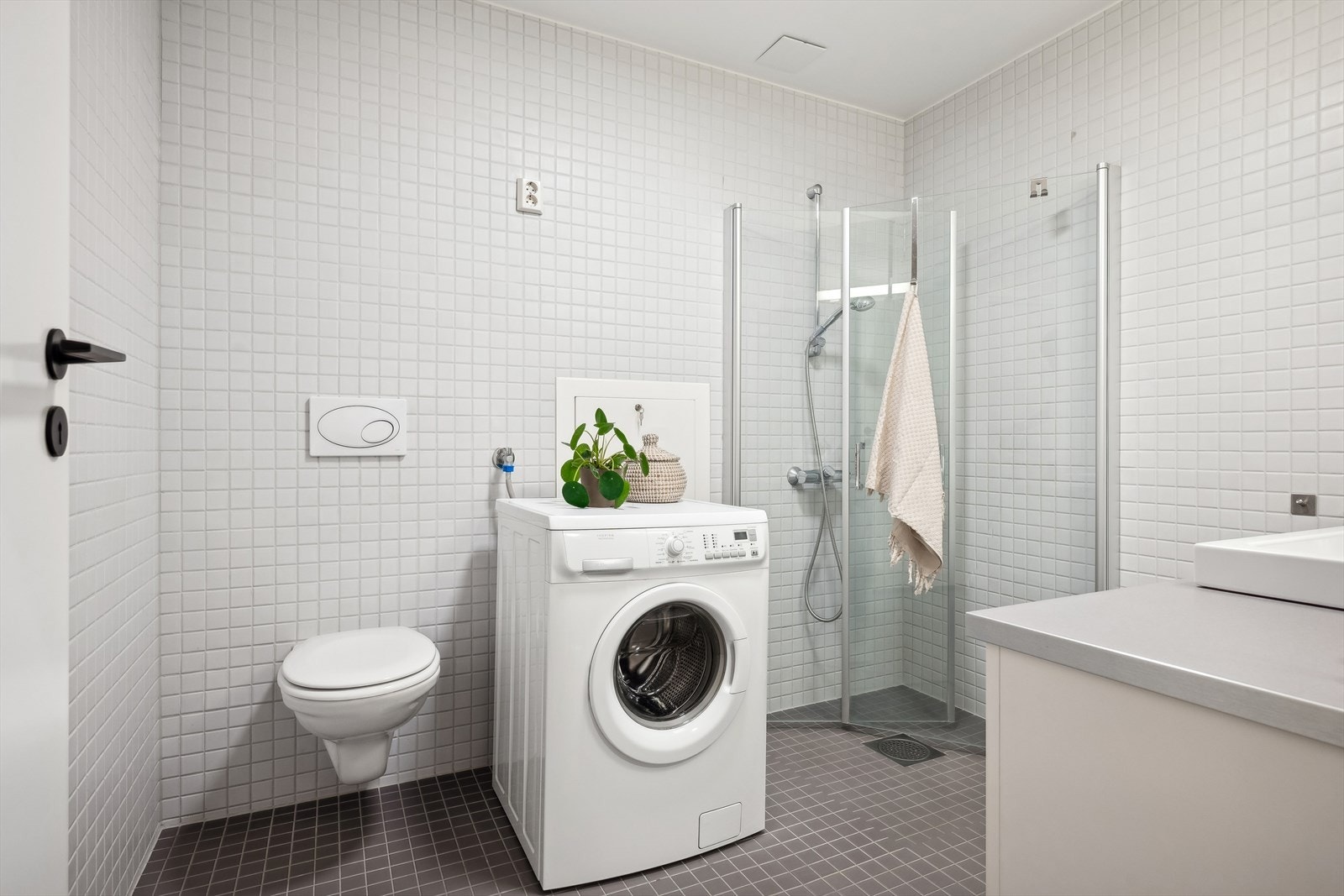 Baderommet inneholder nedfelt servant, underskap, innfliset speil, vegghengt toalett, dusjhjørne med svingbare glassdører og opplegg for vaskemaskin Galleribilde