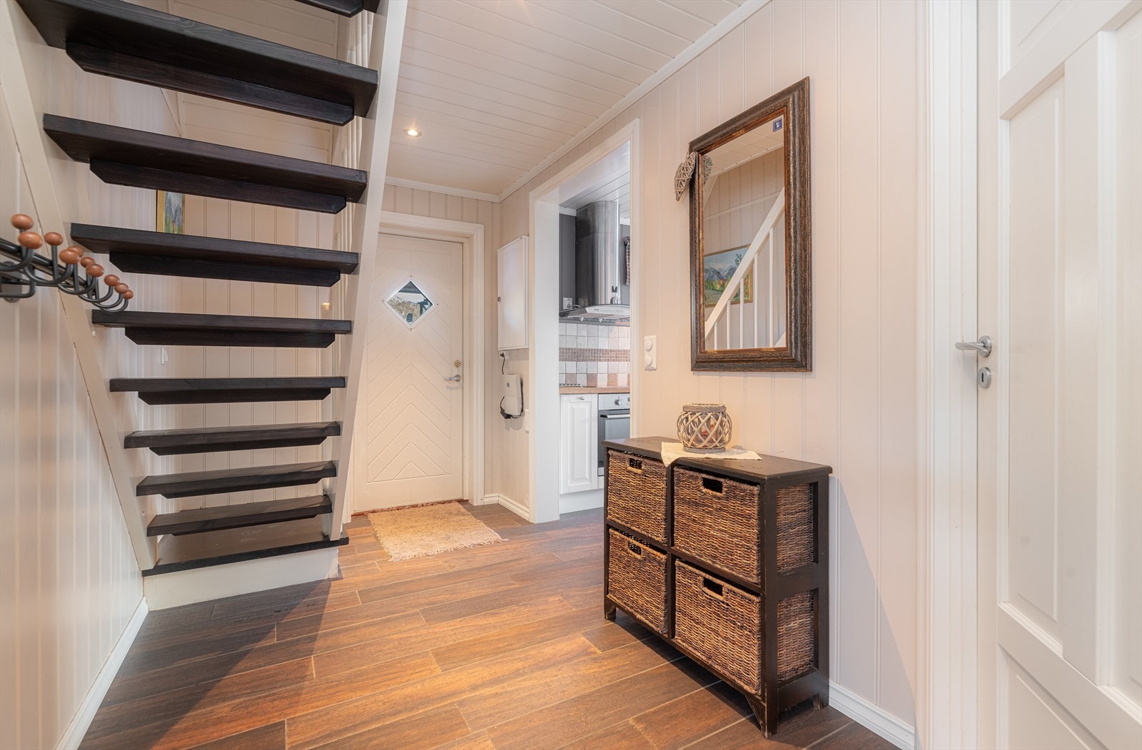 En lys og hyggelig entré ønsker velkommen inn i hytta fra takoverbygget inngangsparti. På innsiden av døren er hyttas sikringsskap innfelt i vegg. Det er malt tretrapp opp til hyttas loftetasje fra gangen, og fin plass til heng av yttertøy. Galleribilde