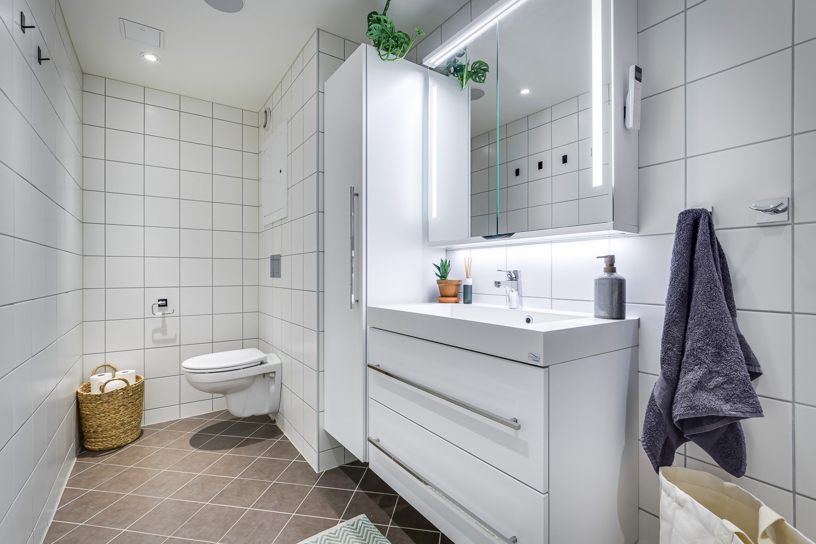 Lys baderomsinnredning med vegghengt servantskap med skuffer, speilskap med belysning og vegghengt skap. Baderommet er også utstyrt med dusjhjørne med innfellbare glassdører og vegghengt toalett. Galleribilde