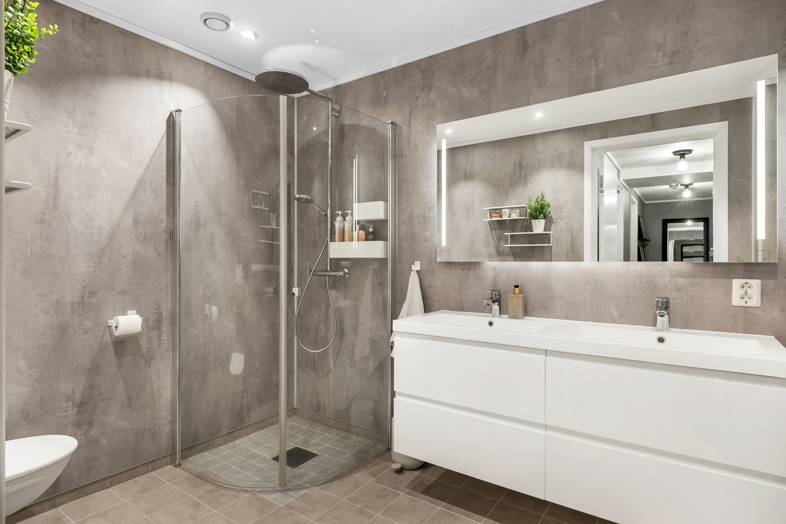 Stort bad med baderomsplater på veggane og flislagt golv. Utstyrt med vegghengt toalett, dusjhjørne og dobbel servant. Galleribilde