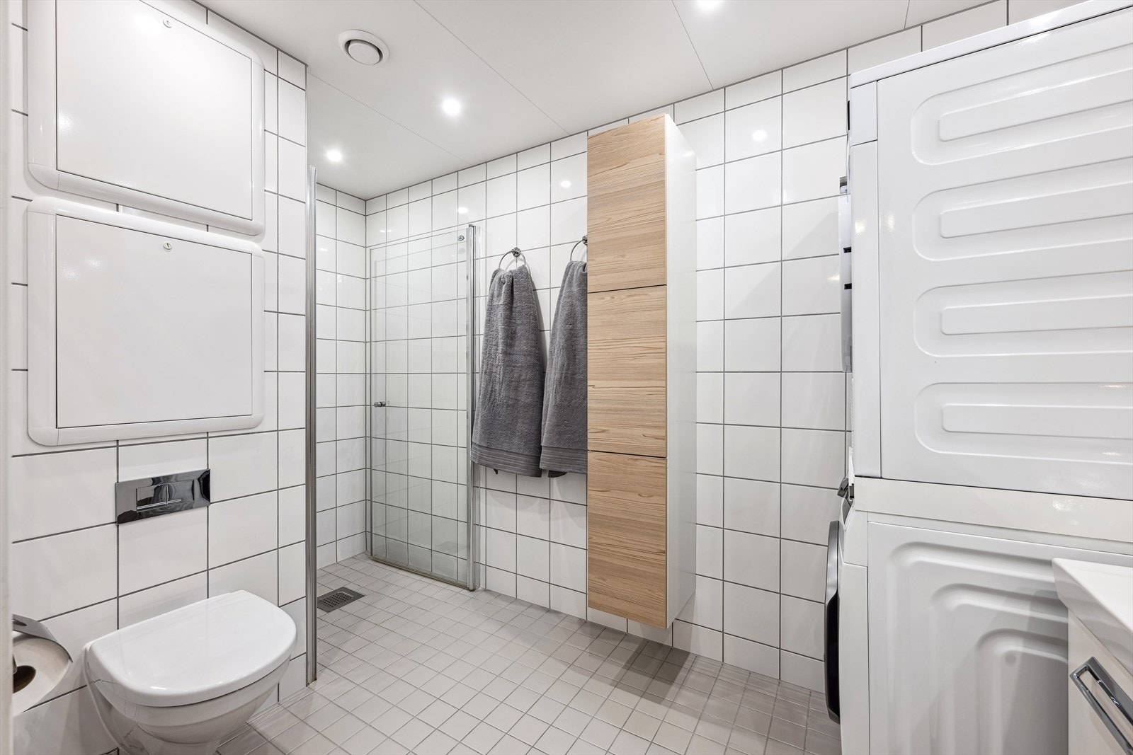 Pent flislagt bad med praktisk innredning med vask, vegghengt wc, dusjnisje, garderobeskap og opplegg for vaskemaskin. Galleribilde