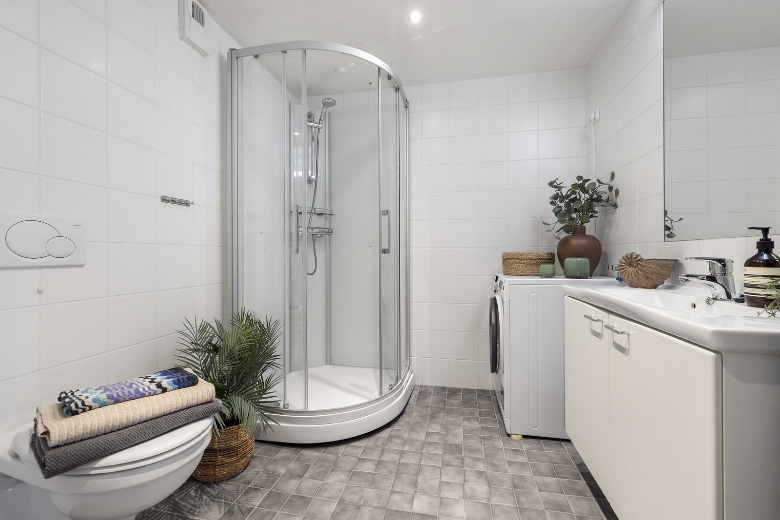 Flislagt baderom med varmekabler og innredning. Badet er utstyrt med servant på benkeskap med speil, vegghengt toalett og dusjhjørne med herdet glass. Galleribilde